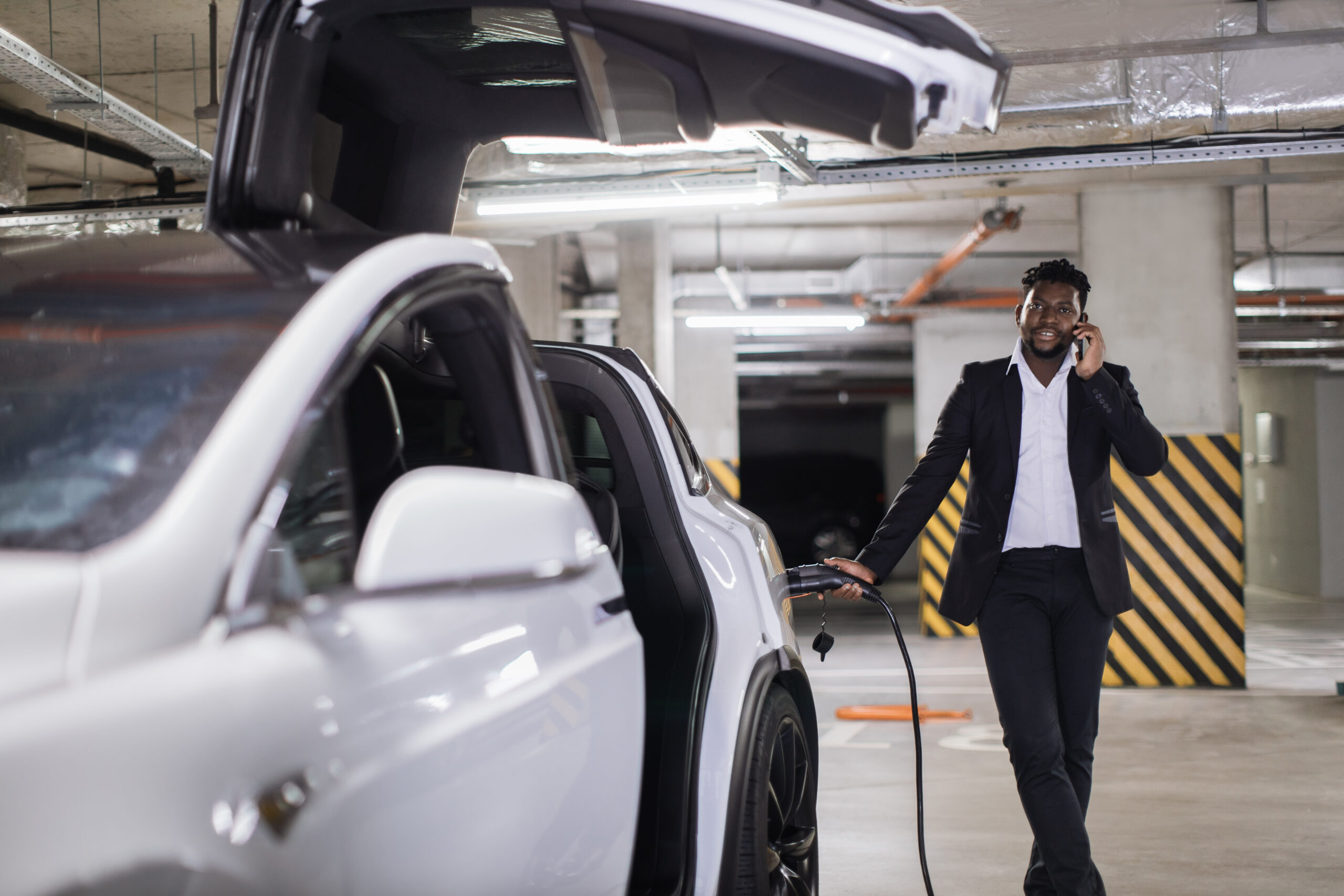 Middle-aged male holding electric connector in battery-drive vehicle while leading phone talk in below-ground car park. Well-organized african american man using time of charging session for business.